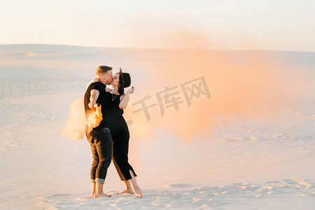 男孩和一个女孩在黑色的衣服拥抱和运行在白色的沙滩上与橙色的烟雾