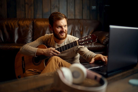 人在耳机演奏吉他听看笔记本电脑屏幕。在线音乐课程男人玩吉他听看笔记本电脑