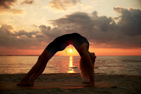 健身、运动和健康的生活方式概念—妇女做瑜伽桥姿势在日落的海滩。妇女做瑜伽桥姿势在海滩