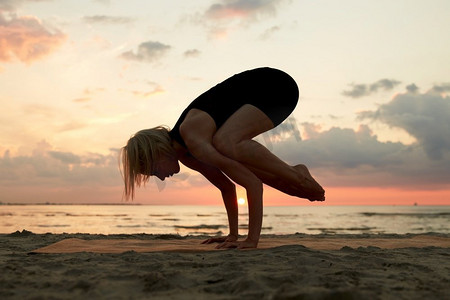 健身、运动和健康的生活方式概念—妇女做瑜伽乌鸦姿势在日落的海滩。妇女做瑜伽乌鸦姿势在海滩上日落