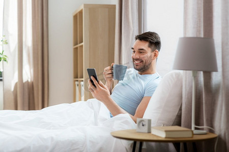 人，睡觉时间和休息的概念-快乐的微笑着的男人，在家里的床上喝着智能手机喝咖啡。家里床上躺着智能手机的快乐男士