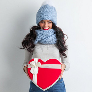 年轻的女孩穿着蓝色编织冬天帽子和围巾与心形礼品盒在白色背景。女孩与心形盒子