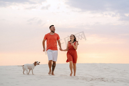 穿着橙色衣服的年轻夫妇带着狗在沙漠中的白沙