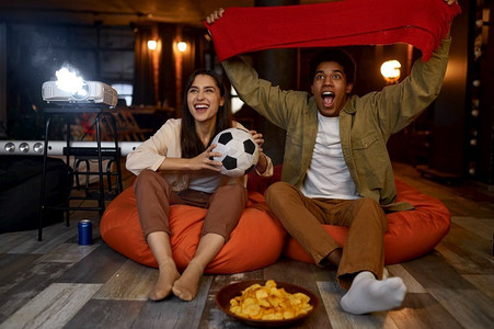 夫妇足球迷观看足球比赛在家庭电视放映机欢呼最喜爱的球队。夫妇观看足球比赛在家庭投影仪