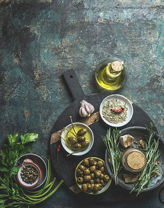 黑暗摄影照片_有橄榄、橄榄油、山柑、盐、大蒜和迷迭香的食物背景在乡村厨房桌子上。地中海美食烹饪俯视图。