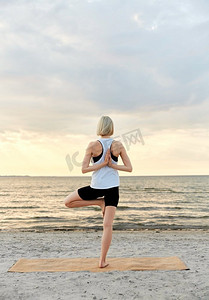 健身，运动，和健康的生活方式概念—妇女做瑜伽树姿势在海滩超过日落。妇女做瑜伽树姿势在海滩在日落