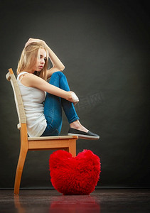 心碎的爱情概念。悲伤的不愉快的妇女坐在椅子红色的心枕头在地板黑暗的背景