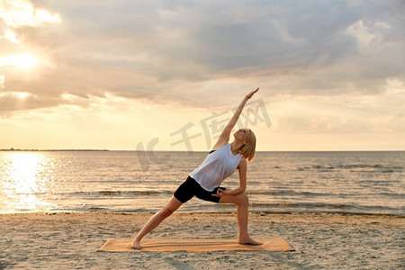健身、运动和健康的生活方式概念—妇女做瑜伽三角姿势在日落的海滩。妇女做瑜伽三角姿势在海滩