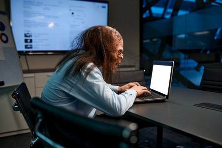 工作在膝上型计算机的僵尸商人在晚上办公室会议室。僵尸商人在办公室工作的笔记本电脑