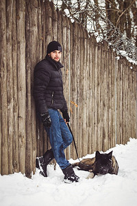 秋摄影照片_一名穿着夹克、戴着针织帽的男子带着一只美国秋田狗穿过雪地森林
