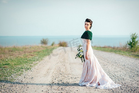新娘手持一束鲜花，身穿象牙色连衣裙，头戴针织围巾，走在延伸至远处的道路上