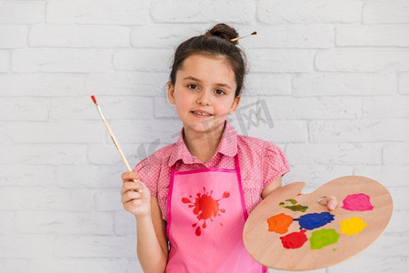 微笑的肖像画女孩站在白色砖墙上，拿着画笔彩色调色板