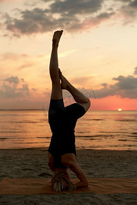 健身、运动和健康的生活方式理念-日落后在海滩上做瑜伽倒立。一名女子在海滩上做瑜伽倒立