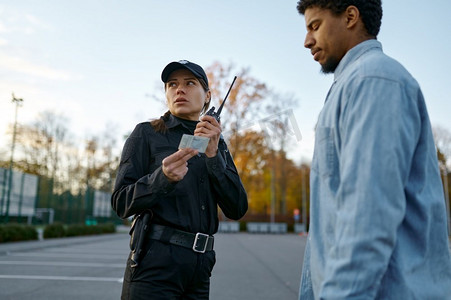 身穿黑色制服的女警察检查男性路人身份证件，对着对讲机说话。女警察检查男性路人身份证件