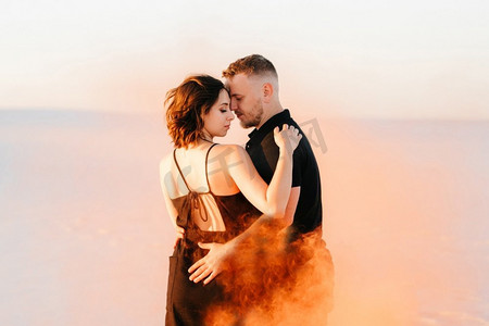 水拥抱摄影照片_男孩和一个女孩在黑色的衣服拥抱和运行在白色的沙滩上与橙色的烟雾