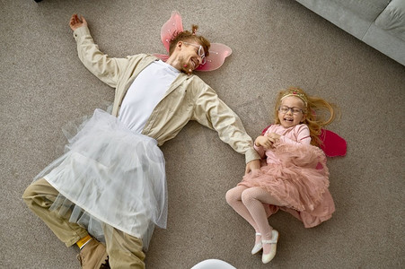 打扮女儿摄影照片_滑稽的穿着父亲和美丽的小女儿微笑的顶视图躺在地板上在家里。有趣的穿着父亲和女儿躺在地板上