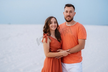 衣服裙子摄影照片_年轻夫妇橙色衣服与狗在沙漠白色的沙滩