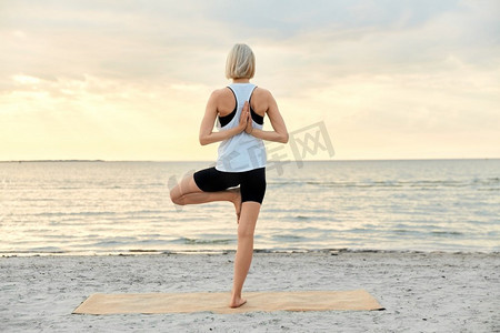 健身，运动，和健康的生活方式概念—妇女做瑜伽树姿势在海滩超过日落。妇女做瑜伽树姿势在海滩在日落