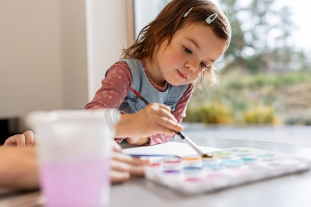 水彩画笔摄影照片_童年，爱好和休闲概念—小女孩在家里用颜色和画笔画画。女婴与颜色和画笔绘画在家里