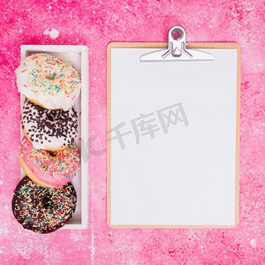 不同类型的甜甜圈白色长方形盒子，靠近剪贴板，粉色背景下有白纸