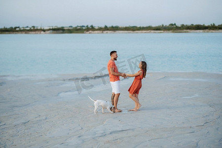 沙滩衣服摄影照片_一对穿着橙色衣服的年轻夫妇带着狗在空荡荡的沙滩上