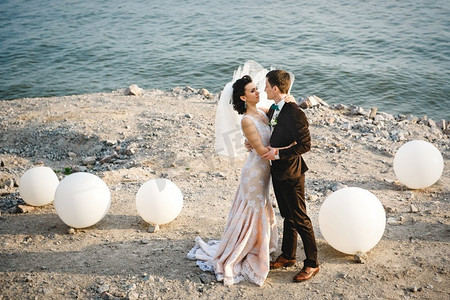 新娘海边摄影照片_穿着棕色西装的新郎和穿着象牙色连衣裙的新娘在多岩石的海边