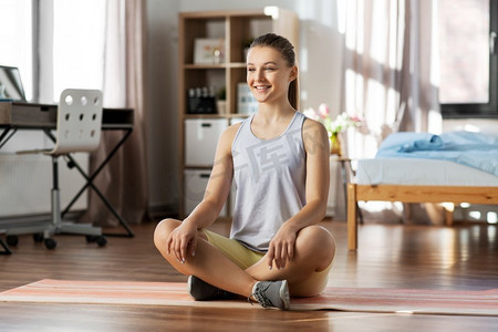 青少年瑜伽摄影照片_运动，健身和健康的生活方式概念—微笑的少女坐在瑜伽垫在家里。微笑的十几岁的女孩坐在瑜伽垫在家