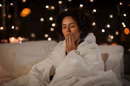 人,就寝时间和休息概念-疲倦困倦的女人裹着毯子,晚上坐在床上打哈欠.