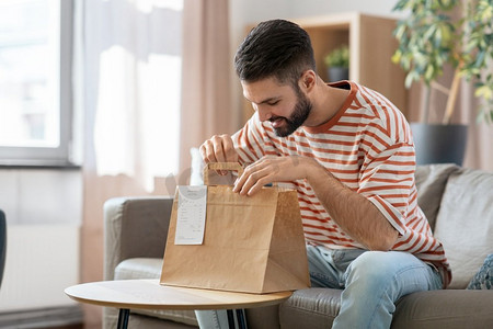 消费，吃和人的概念—微笑的男人打开外卖食品在纸袋在家里。微笑的男人在家里打开外卖食品