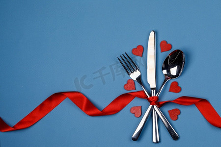 红色叉摄影照片_餐具套与红色丝带和心脏绑在蓝色背景情人节晚餐概念。情人节晚餐餐具套装