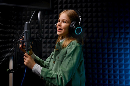 年轻的少女明星歌手戴耳机唱新歌与麦克风在唱片室。年轻的青少年女孩歌手在记录工作室