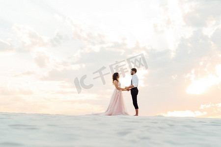 裤子裙子摄影照片_一对年轻的夫妇，一个穿着黑色裤子的男孩和一个穿着粉红色裙子的女孩正沿着沙漠的白色沙滩走着