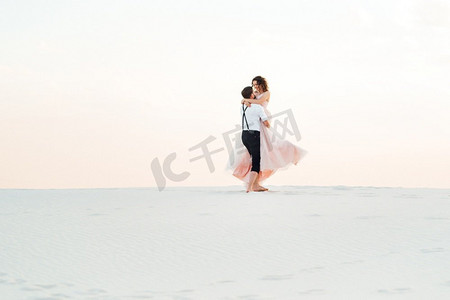 裤子裙子摄影照片_一对年轻的夫妇，一个穿着黑色裤子的男孩和一个穿着粉红色裙子的女孩正沿着沙漠的白色沙滩走着