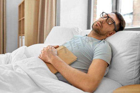 人，就寝时间和休息的概念—戴眼镜的人与书睡在床上在家里早上。男人在眼镜与书睡觉在家里的床上