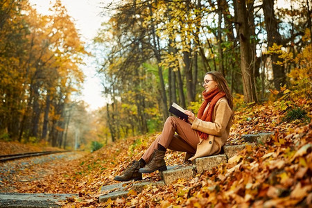 秋天读书摄影照片_坐在楼梯读书在秋天背景的聪明的年轻女子地上有金色的叶子。年轻女子与书在秋天背景