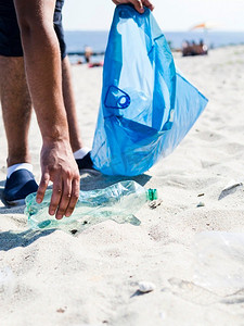 奥林巴斯数码相机。一名男子手持蓝色垃圾袋在海滩边捡垃圾塑料瓶