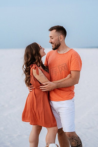夏天衣服摄影照片_穿着橙色衣服的年轻夫妇带着狗在沙漠中的白沙
