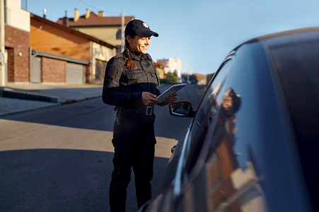 年轻的女警察与平板电脑微笑对站在汽车附近的司机.女交警在路上工作。年轻的女警察与平板电脑站在汽车附近