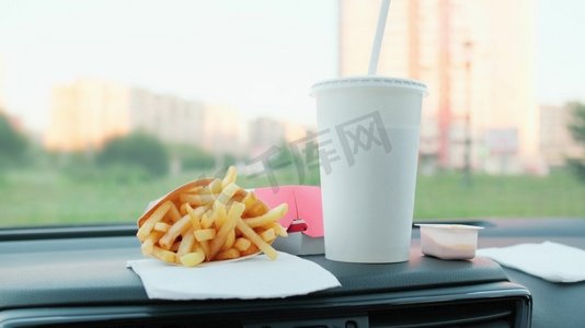 在汽车碰撞时吃快餐。汉堡，炸薯条和饮料在车上用吸管。模糊的城市风景背景。汉堡，薯条和饮料在车里。