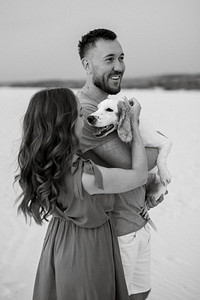 夏天衣服摄影照片_穿着橙色衣服的年轻夫妇带着狗在沙漠中的白沙