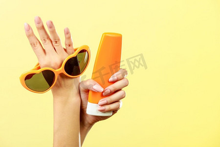 假日夏季时尚眼部护肤概念。妇女拿着手心形太阳镜防晒乳液，黄色背景