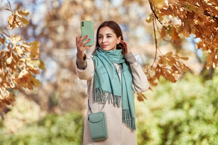 一个年轻漂亮的女人走过秋天的公园。一个女孩拿着智能手机在秋天的森林里拍照