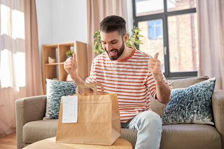 消费，吃和人的概念—微笑的男人打开外卖食品在纸袋在家里。微笑的男人在家里打开外卖食品