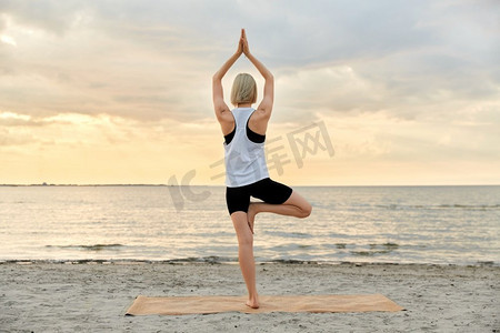 健身、运动和健康生活方式理念--日落时分，女性在海滩上摆瑜伽树姿势。日落时分，一名女子在海滩上摆瑜伽树姿势
