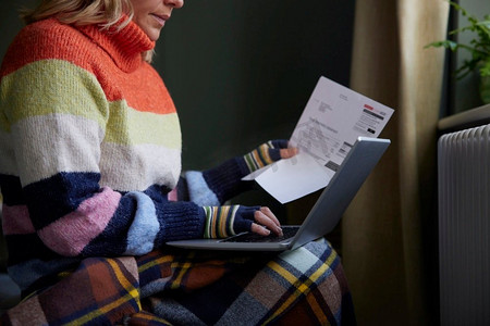 戴手套的女人与笔记本电脑和比尔试图保持温暖的散热器在成本的生活能源危机