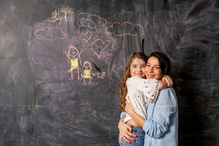 母亲孩子拥抱摄影照片_愉快的母亲女儿拥抱在黑板附近与绘图