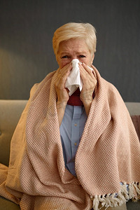 不健康的老妇人感到生病裹在毯子里擦鼻涕成餐巾。不健康的生病的老妇人裹在毯子