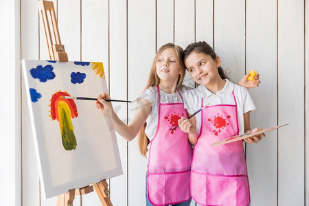 水果画摄影照片_快乐的女孩拿着调色板的手看她的朋友画画布用刷子