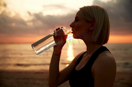 健身，运动，和健康的生活方式概念—妇女饮用水从瓶子在海滩超过日落。妇女从海滩上的瓶子喝水
