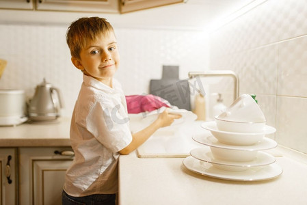 一个戴手套的小男孩在厨房里洗盘子。宝宝在家做家务。年轻妈妈的助手打扫房子’